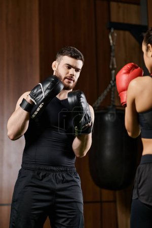 Photo for A male trainer wearing boxing gloves stands next to a brunette sportswoman in active wear during a boxing session in a gym. - Royalty Free Image
