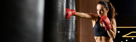 A brunette sportswoman in a black sports bra and red boxing gloves is fiercely boxing in a gym.