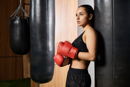 Une sportive brune confiante en tenue active se tient à côté d'un sac de boxe portant des gants de boxe rouges, prête pour une séance d'entraînement intense.