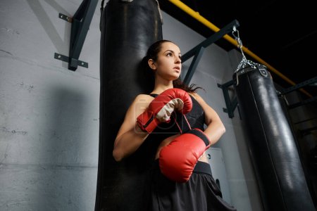 A determined brunette sportswoman wearing red boxing gloves stands confidently next to a punching bag in a gym.