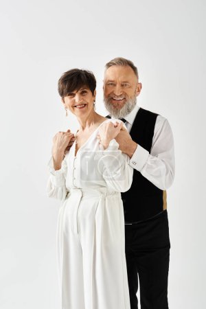A middle aged bride and groom in wedding gowns stand next to each other, celebrating their special day in a studio setting.