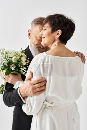Photo for Middle-aged bride and groom in wedding attire hugging tightly while holding flowers, celebrating their special day in a studio setting. - Royalty Free Image