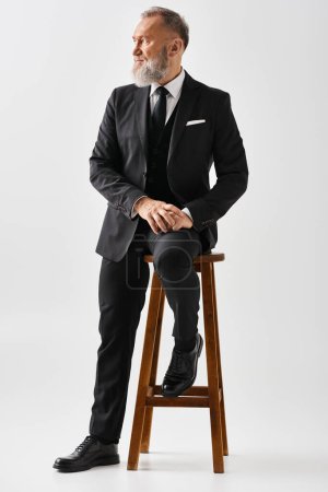 Photo for A middle-aged groom in an elegant suit sits on a stool in a studio setting on his wedding day. - Royalty Free Image