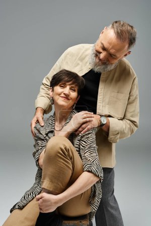 Un homme d'âge moyen soutient une femme sur le dossier d'une chaise dans un cadre élégant de studio, montrant la confiance et le partenariat.