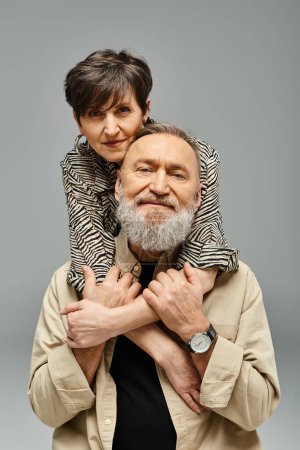 Un homme d'âge moyen en tenue élégante soulève une femme sur ses épaules dans un cadre de studio, montrant l'affection et la force.