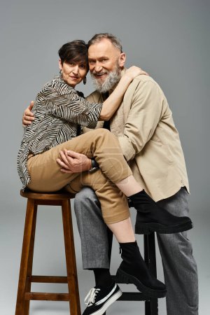 Mann und Frau mittleren Alters in stilvoller Kleidung sitzen auf einem Hocker in einem Studio-Ambiente.