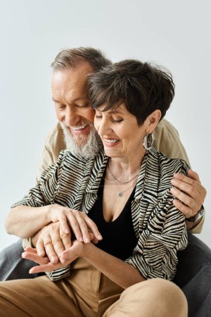 A middle-aged couple dressed stylishly, seated together on a chair, exuding grace and togetherness in a chic studio setting.