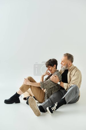A middle-aged couple in stylish attire seated closely together in a studio setting, exuding sophistication and unity.