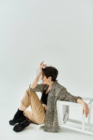 A woman in stylish clothes sits on the ground next to a pristine white chair in a contemplative pose.