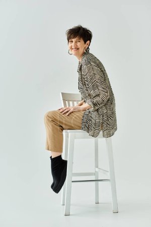 A stylish middle-aged woman with short hair sits on a stool in a studio setting.