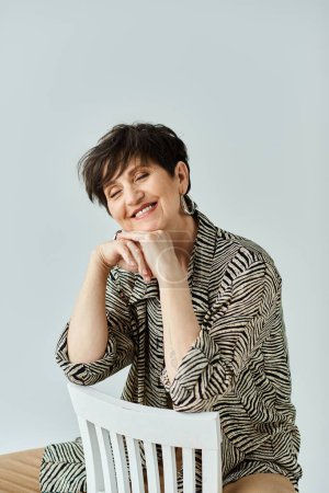 Middle-aged woman with short hair dressed elegantly, sits gracefully on a white chair in a studio setting.