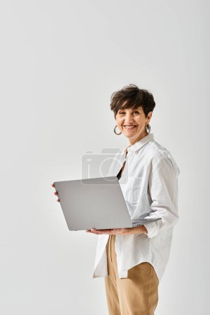 Foto de Una mujer de mediana edad con un atuendo elegante y cabello corto sostiene una computadora portátil en sus manos en un entorno de estudio. - Imagen libre de derechos