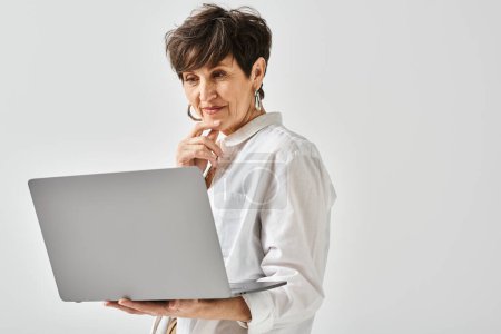 A middle-aged woman in stylish attire confidently holds a laptop computer in her hands in a studio setting.