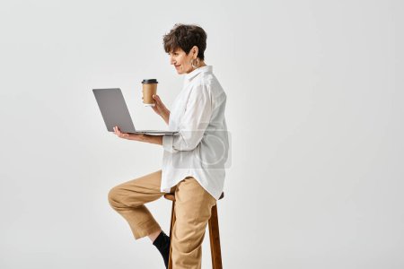 Foto de Una mujer de mediana edad con un atuendo elegante se sienta en un taburete, trabajando en una computadora portátil en un entorno de estudio. - Imagen libre de derechos