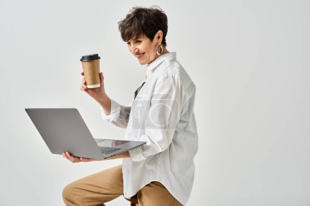 A middle-aged woman in stylish attire holding a cup of coffee and working on her laptop in a studio setting.