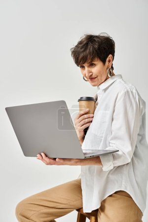 Téléchargez les photos : Une femme d'âge moyen élégamment habillée avec des cheveux courts s'assoit sur un tabouret, multitâche avec une tasse de café dans une main et un ordinateur portable dans l'autre. - en image libre de droit