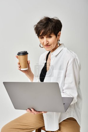 Multitarea de mujer de mediana edad, sosteniendo una taza de café y una computadora portátil en un elegante entorno de estudio.