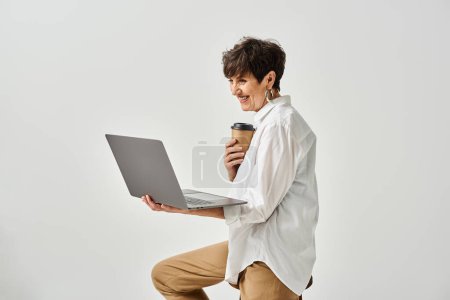 A middle-aged woman sitting on a stool, holding a laptop in a stylish studio setting.