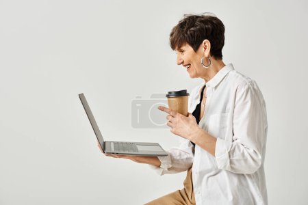 A middle aged woman in stylish attire holding a cup of coffee while working on a laptop in a studio setting.
