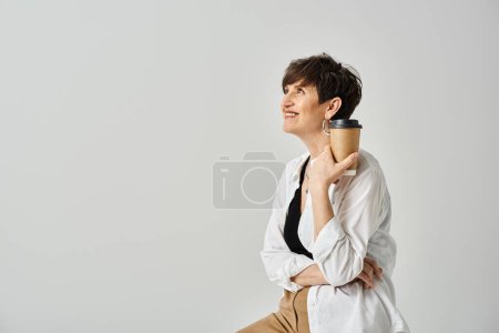 A stylish, middle-aged woman sits on a stool, delicately holding a cup of coffee.