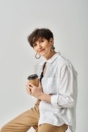 Foto de Una mujer de mediana edad con el pelo corto se sienta en un taburete, vestido con estilo, sosteniendo una taza de café en un momento sereno. - Imagen libre de derechos