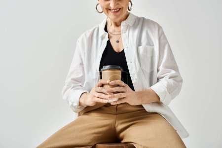 A middle-aged woman in stylish attire sits on a stool, serenely holding a cup of coffee.