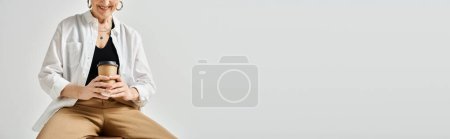 Téléchargez les photos : Femme d'âge moyen avec les cheveux courts dans une tenue élégante, assis sur un tabouret, absorbé dans son téléphone portable. - en image libre de droit