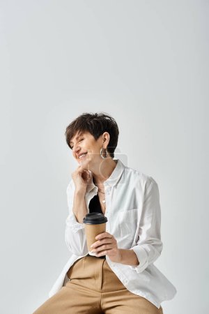 Una mujer de mediana edad, elegantemente vestida, con el pelo corto se sienta en un taburete, sosteniendo delicadamente una taza de café, perdida en el pensamiento.