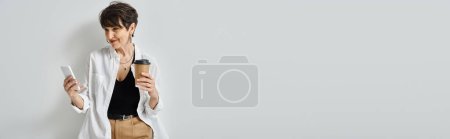 Photo for A middle aged woman with short hair holds a cell phone and a cup of coffee in a stylish studio setting. - Royalty Free Image