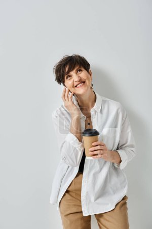 A middle-aged woman with short hair, dressed stylishly, multitasking by holding a cup of coffee and chatting on her cellphone.