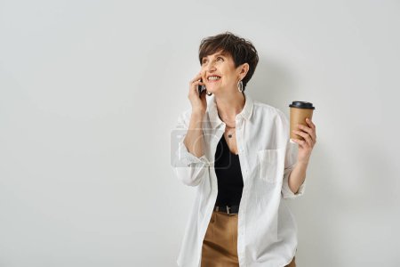 Stilvolle Frau mittleren Alters mit kurzen Haaren, Multitasking, hält eine Tasse Kaffee in der Hand, während sie mit dem Handy spricht.