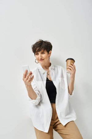 A stylish middle-aged woman with short hair is holding a cup of coffee in one hand and a cell phone in the other.