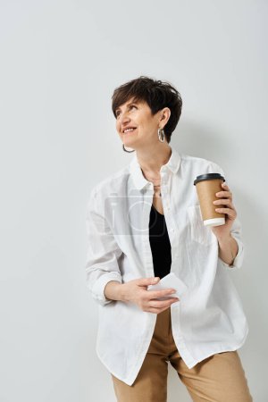 Foto de Una mujer elegante de mediana edad con sonrisas de pelo corto mientras sostiene una taza de café en un ambiente acogedor estudio. - Imagen libre de derechos