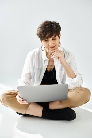 Foto de Una mujer de mediana edad elegantemente vestida con el pelo corto se sienta en el suelo, centrada en un ordenador portátil. - Imagen libre de derechos