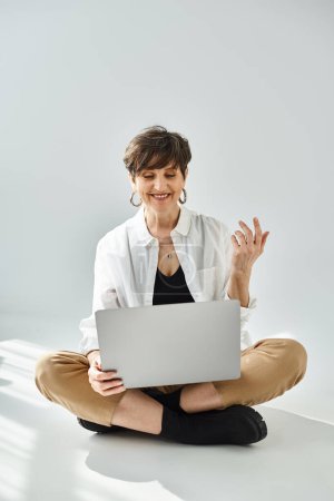 Téléchargez les photos : Une femme d'âge moyen aux cheveux courts est assise sur le sol, intensément concentrée sur son ordinateur portable dans un cadre de studio élégant. - en image libre de droit