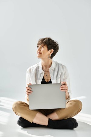 Una mujer de mediana edad con el pelo corto vestido con estilo, sentado en el suelo y trabajando en un ordenador portátil en un entorno de estudio.