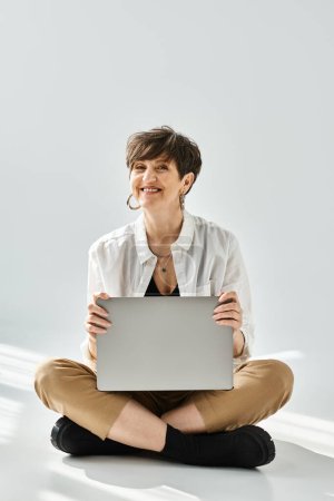 Téléchargez les photos : Une femme d'âge moyen élégamment habillée aux cheveux courts est assise sur le sol, travaillant sur un ordinateur portable dans un cadre de studio. - en image libre de droit