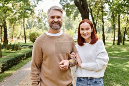 A man and woman in casual attire stand close in a peaceful park setting.