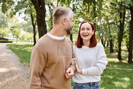 A man and a woman in casual attire stroll through a lush park.