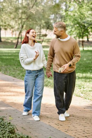 A man and woman in casual attire walk together on a sidewalk in a park.