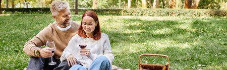 Erwachsene Männer und Frauen sitzen auf einer Decke und halten Weingläser in einem Park.