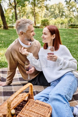 Ein Mann und eine Frau sitzen auf einer Decke in einem Park und halten Weingläser.