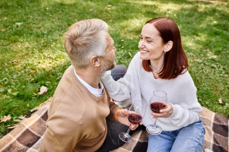 A man and a woman sit on a blanket, holding wine glasses in a park.