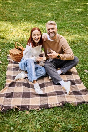 Un couple assis sur une couverture, tenant des verres à vin dans un parc.