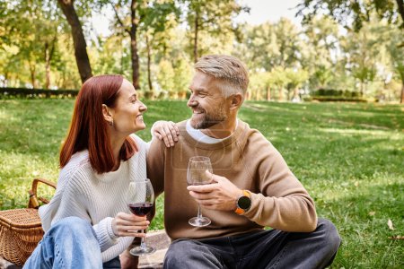 A couple sits on a blanket in a park holding wine glasses.