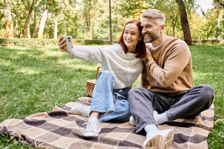 Mann und Frau sitzen auf Decke im Park und halten Moment mit Selfie fest.