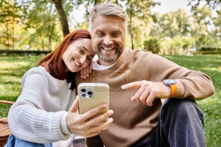 Téléchargez les photos : Un couple en tenue décontractée prend romantique un selfie avec un téléphone portable dans le parc. - en image libre de droit