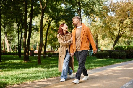 A man and woman in casual attire walk down a peaceful path in a lush park.
