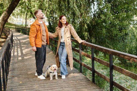 A couple in casual attire walks their dog on a bridge in the park.