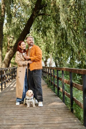 Foto de Un hombre y una mujer con un perro de pie en un puente, disfrutando de un paseo pausado. - Imagen libre de derechos